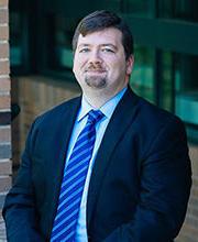 Professor Nicholas Ganson smiling with short brown hair, mustache and goatee wearing a navy blue blazer over a light blue button down shirt and blue striped tie