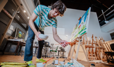 Mason Terra holds a brush and bowl as he paints on a canvas 